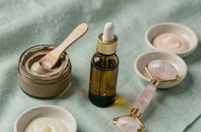 Mise en scène flat lay de produits de soins de beauté haut de gamme sur lin vert sauge incluant sérum, masque à l'argile et rouleau en quartz rose